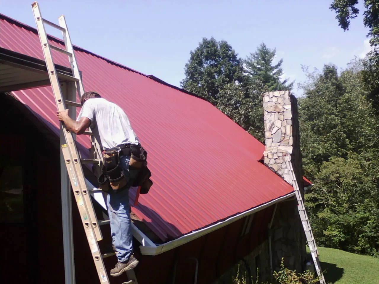 Roofer on a ladder beside a red metal roof and stone chimney — Soffit & Fascia Repair by Roofing Brothers in Exton